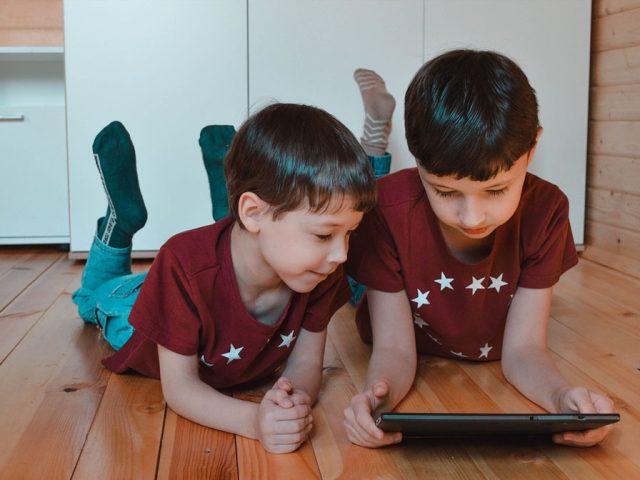 Two young boys laying on their stomachs, propped up on their elbows and watching a tablet. Image:image Pixabay/Vika Glitter